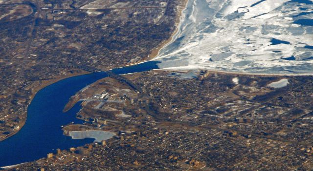Aerial view of Highway 402 passing through Sarnia. The Blue Water Bridge is visible, and Lake Huron can be seen at the top of the image, still frozen. Image: WikiCommons. 
