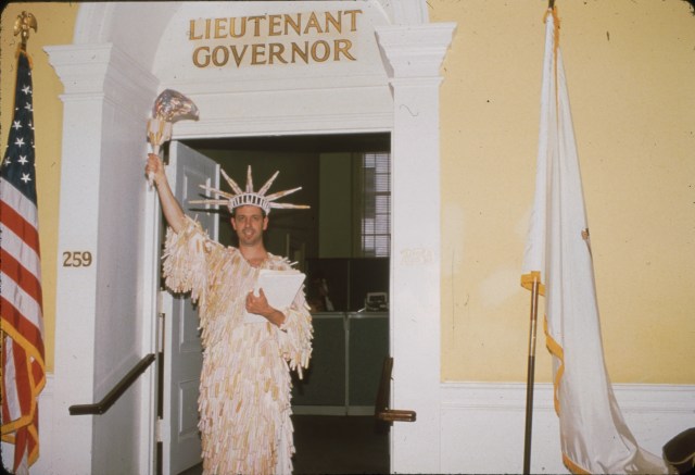 A person in a white Statue of liberty costume stands in a doorway, flanked by an American flag